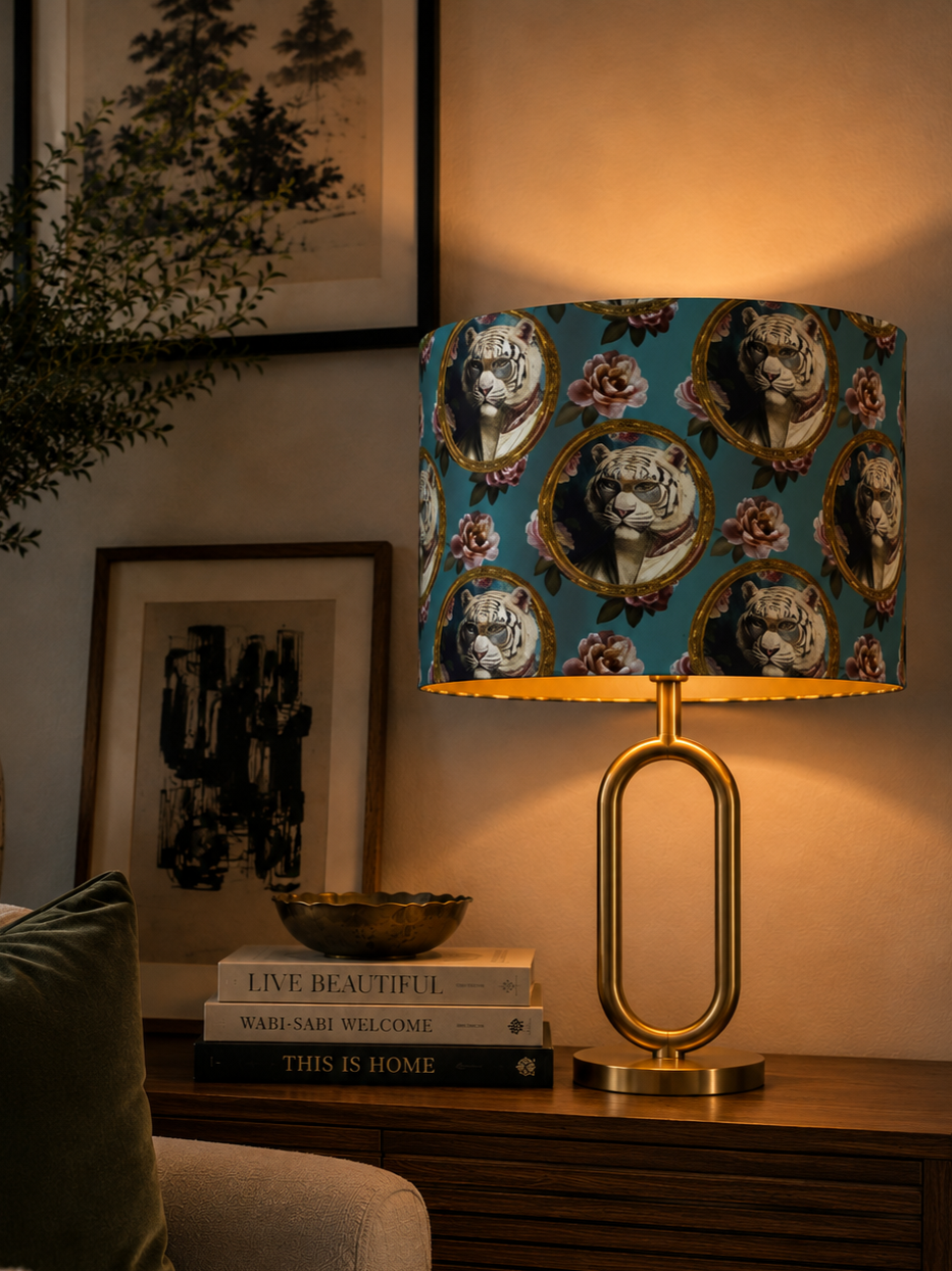 A decorative table lamp with a tiger pattern lampshade on a wooden surface, next to stacked books and framed artwork on the wall.