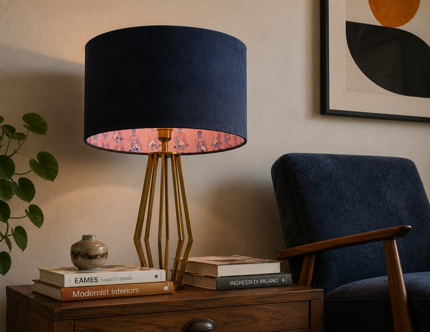 A room corner with a navy blue lamp that has a gold geometric base, a wooden side table, a dark blue armchair, and a framed abstract art print on the wall. The table has a stack of books and a small ceramic vase, with a potted plant beside it.