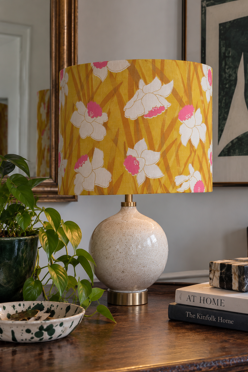Table lamp with a floral yellow lampshade, a potted green plant, a black and white speckled dish, and stacked books on a wooden surface, with a large mirror and framed artwork in the background.