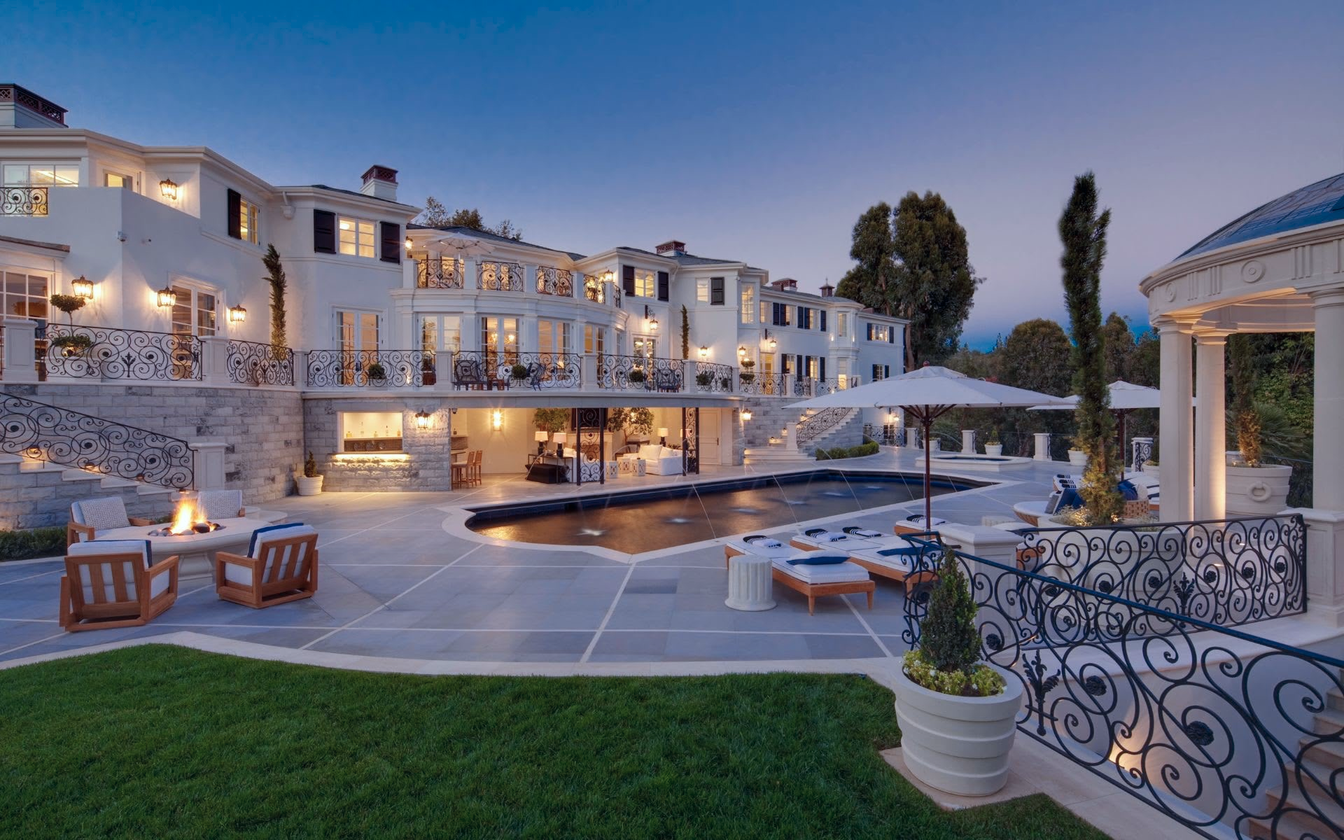 Luxury mansion with outdoor pool area, lounge chairs, umbrellas, fire pit, and elegant architecture at dusk.
