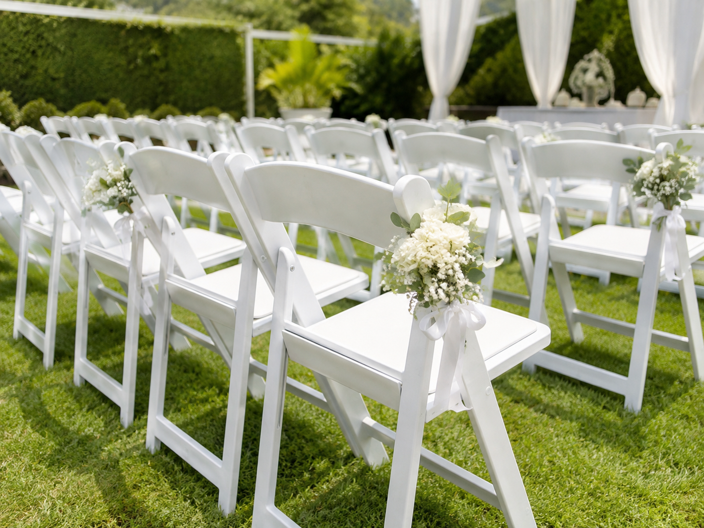 White folding chairs decorated with white floral arrangements and ribbons set up outdoors on a grassy area for a wedding ceremony.