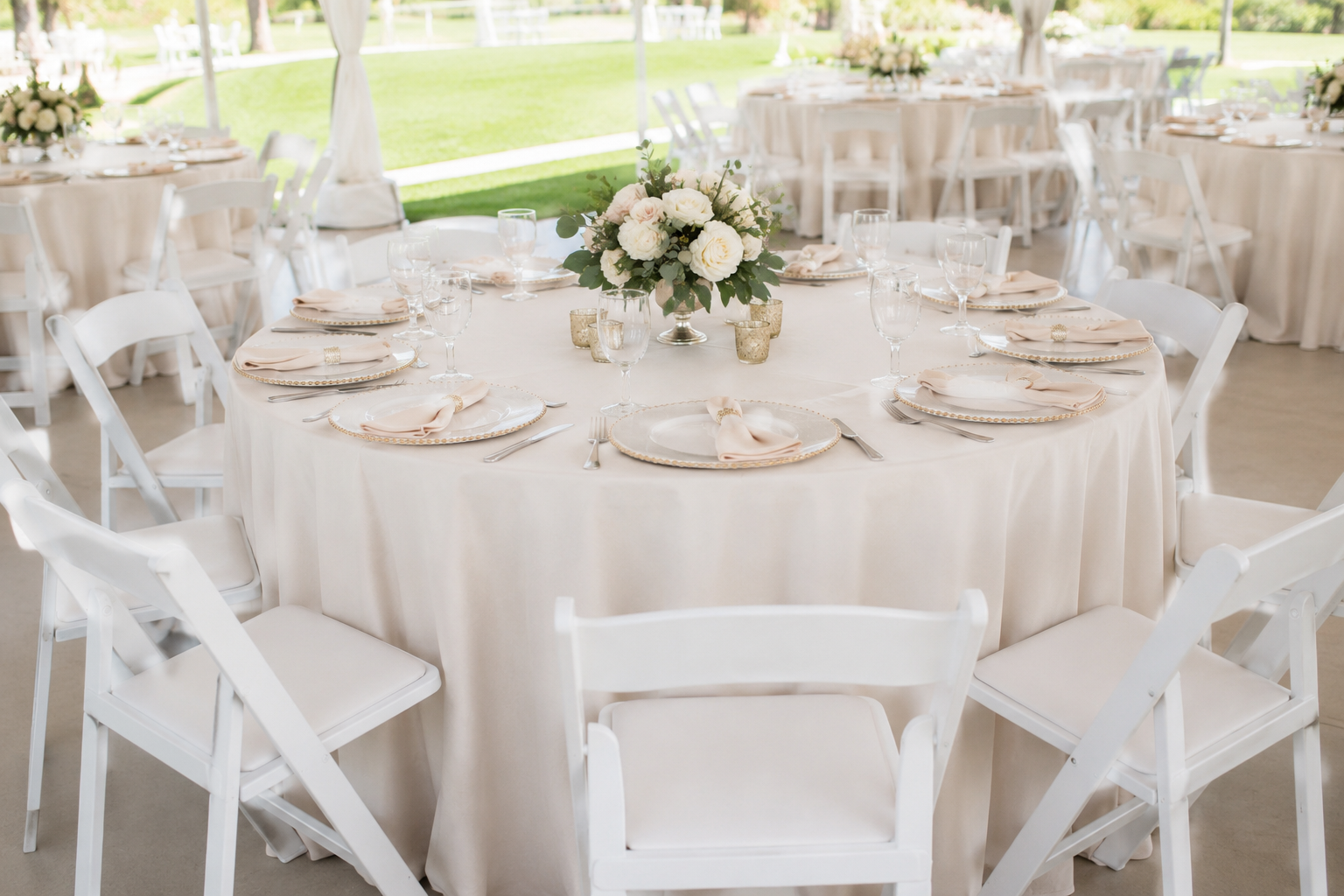 Elegant outdoor wedding reception setup with a round table draped in a white tablecloth, decorated with a floral centerpiece of white roses and greenery, set with white plates, blush napkins, silverware, wine glasses, and small gold votive candle holders, surrounded by white chairs.
