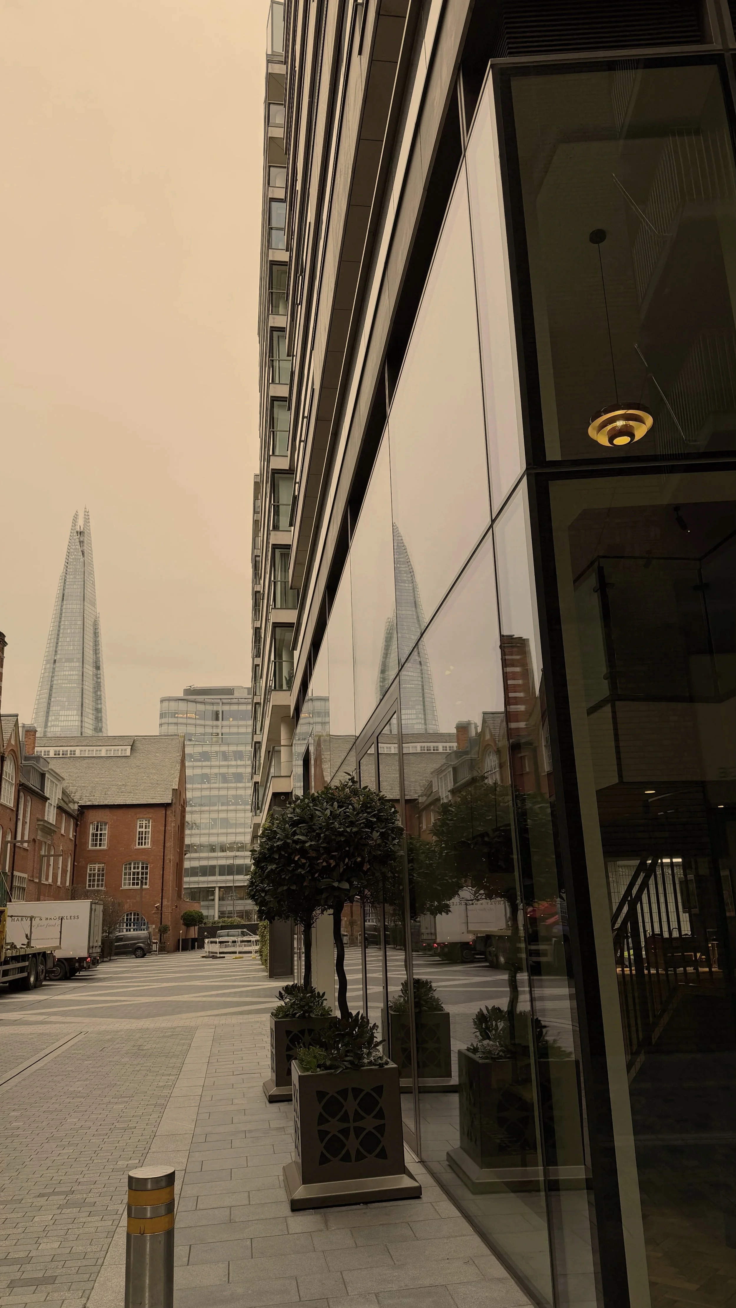 City street with modern glass building reflecting older buildings, including the Shard skyscraper, with potted plants and trees along the sidewalk.