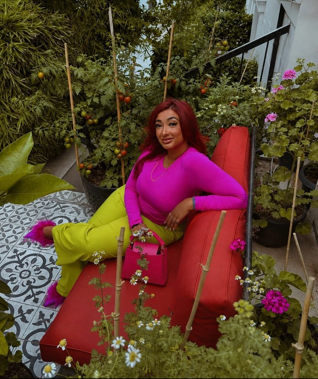 A woman with red hair sitting on a red outdoor cushioned sofa surrounded by potted plants, including tomato plants and flowering plants, on a patterned tile patio.