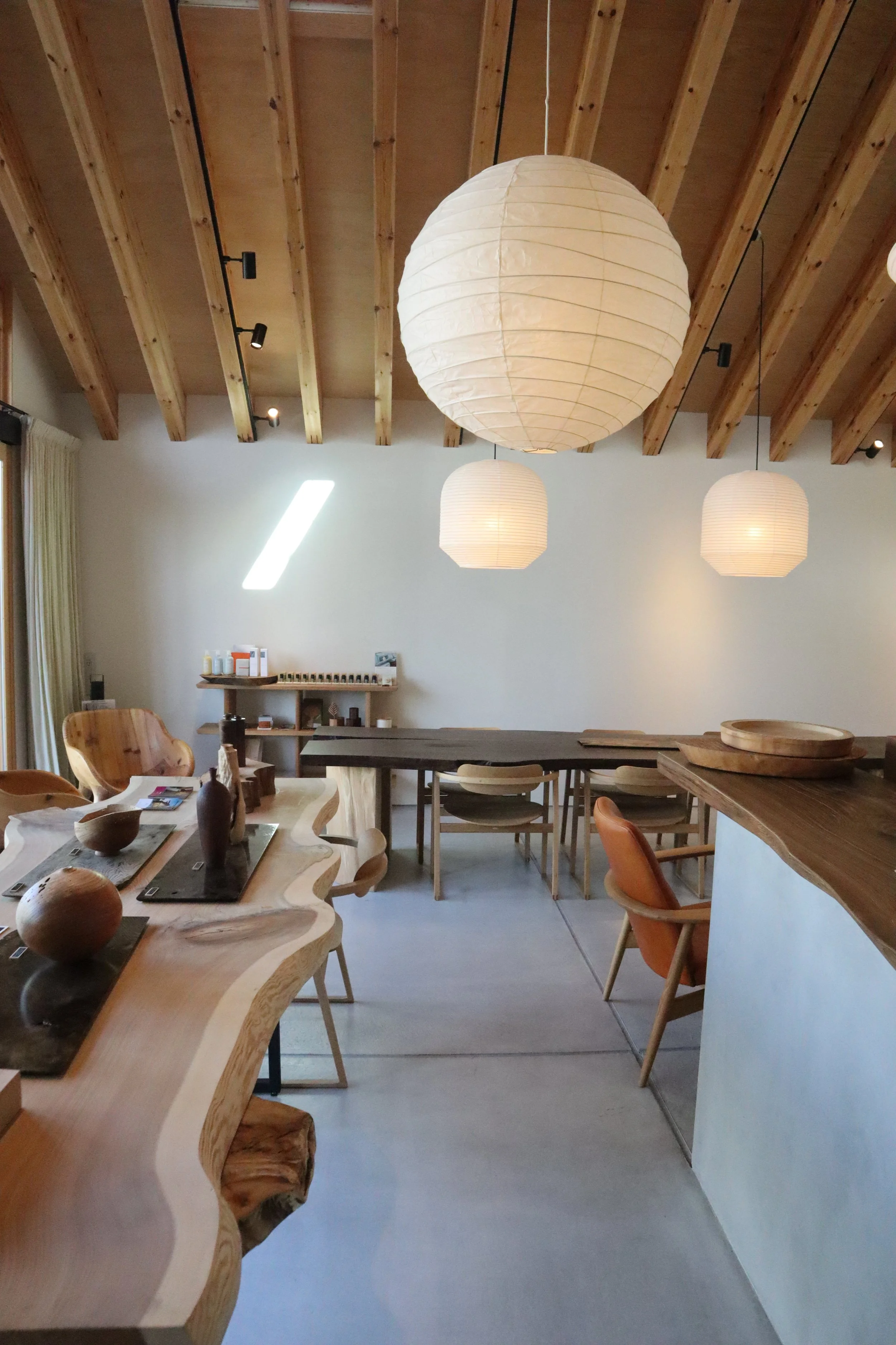 Interior of a modern room with wooden beams on the ceiling, hanging paper lantern lights, and a variety of wooden and upholstered chairs around a long table.
