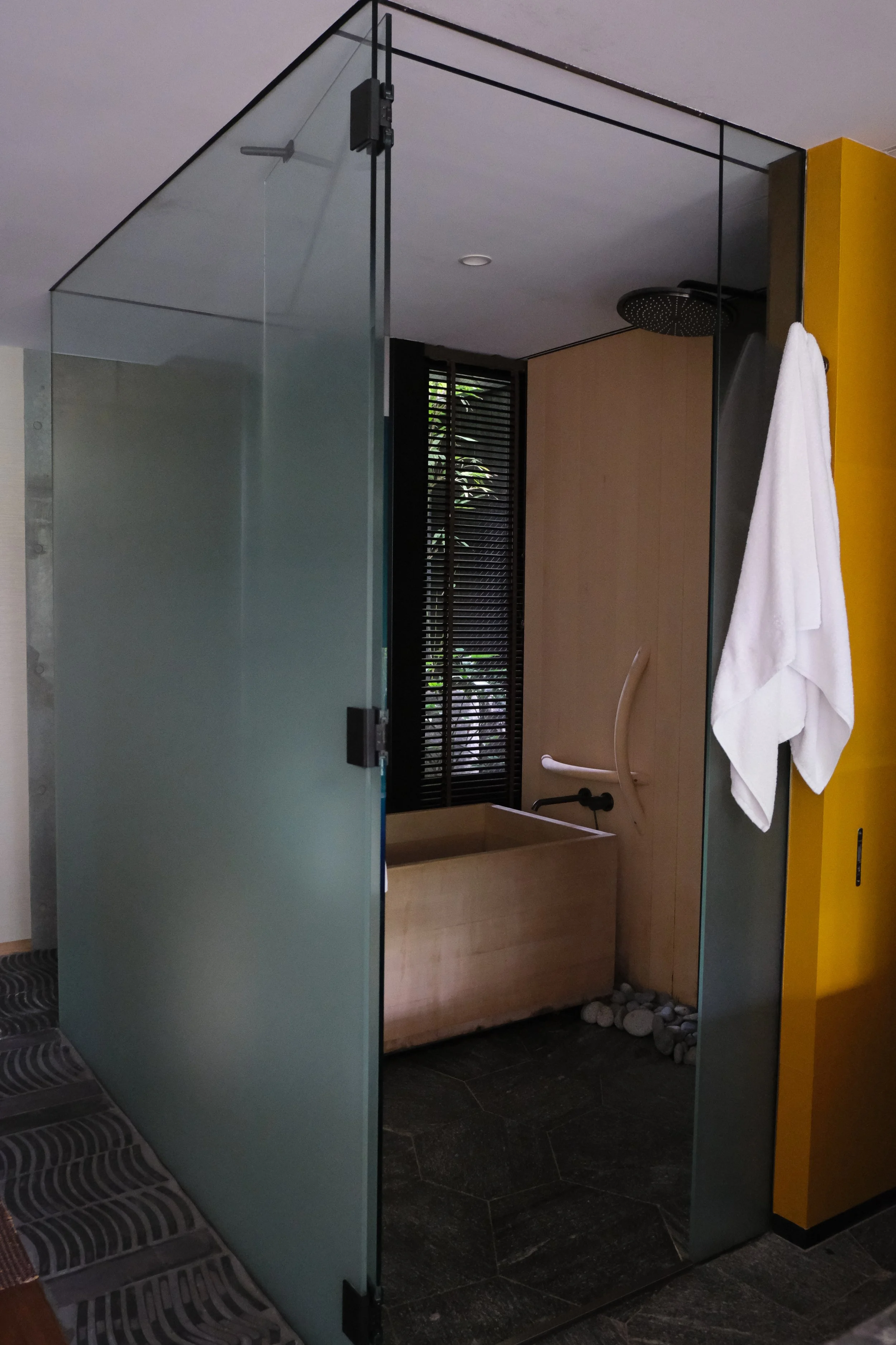 A modern outdoor shower with a wooden base and a rain shower head, enclosed by glass panels, with a white towel hanging on the right side.