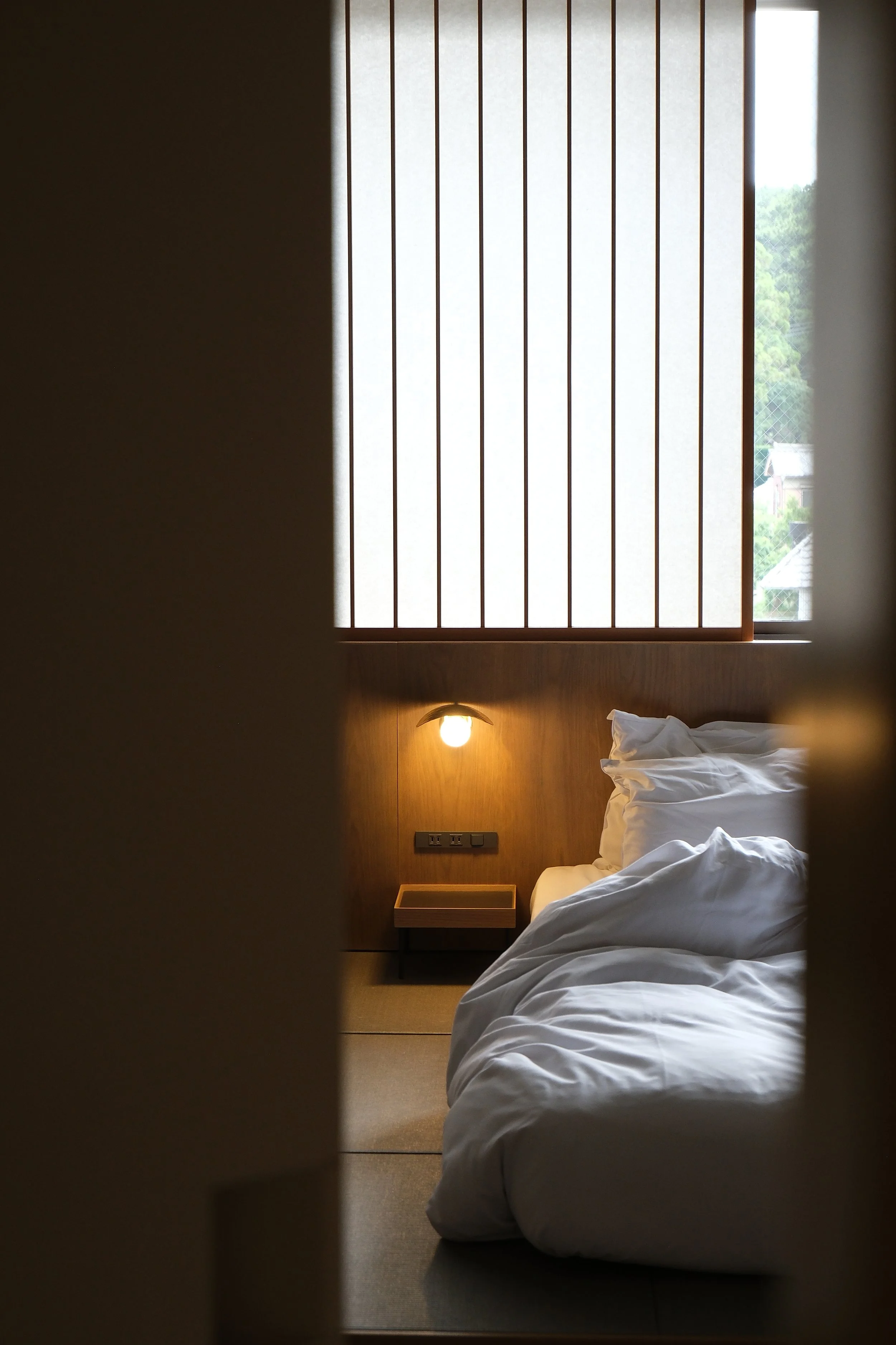 A partially opened hotel room door reveals a bed with white linens, a wooden headboard, a small bedside table, a wall-mounted lamp, and a large window with vertical blinds allowing natural light into the room.