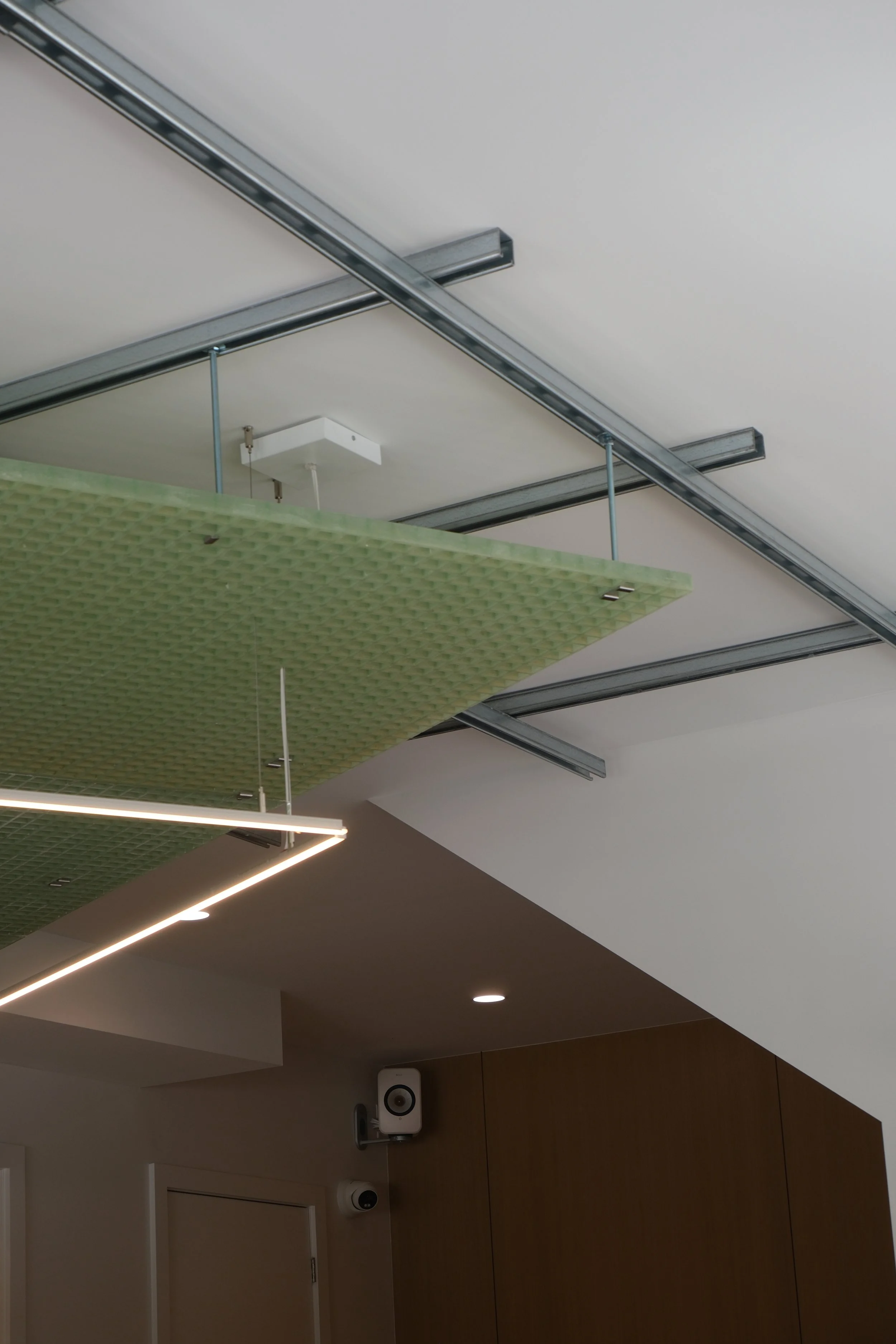 Ceiling with metallic support beams, a green textured panel hanging from the ceiling, and a modern LED light fixture. Also visible are two speakers and a white speaker or device mounted on the wall.