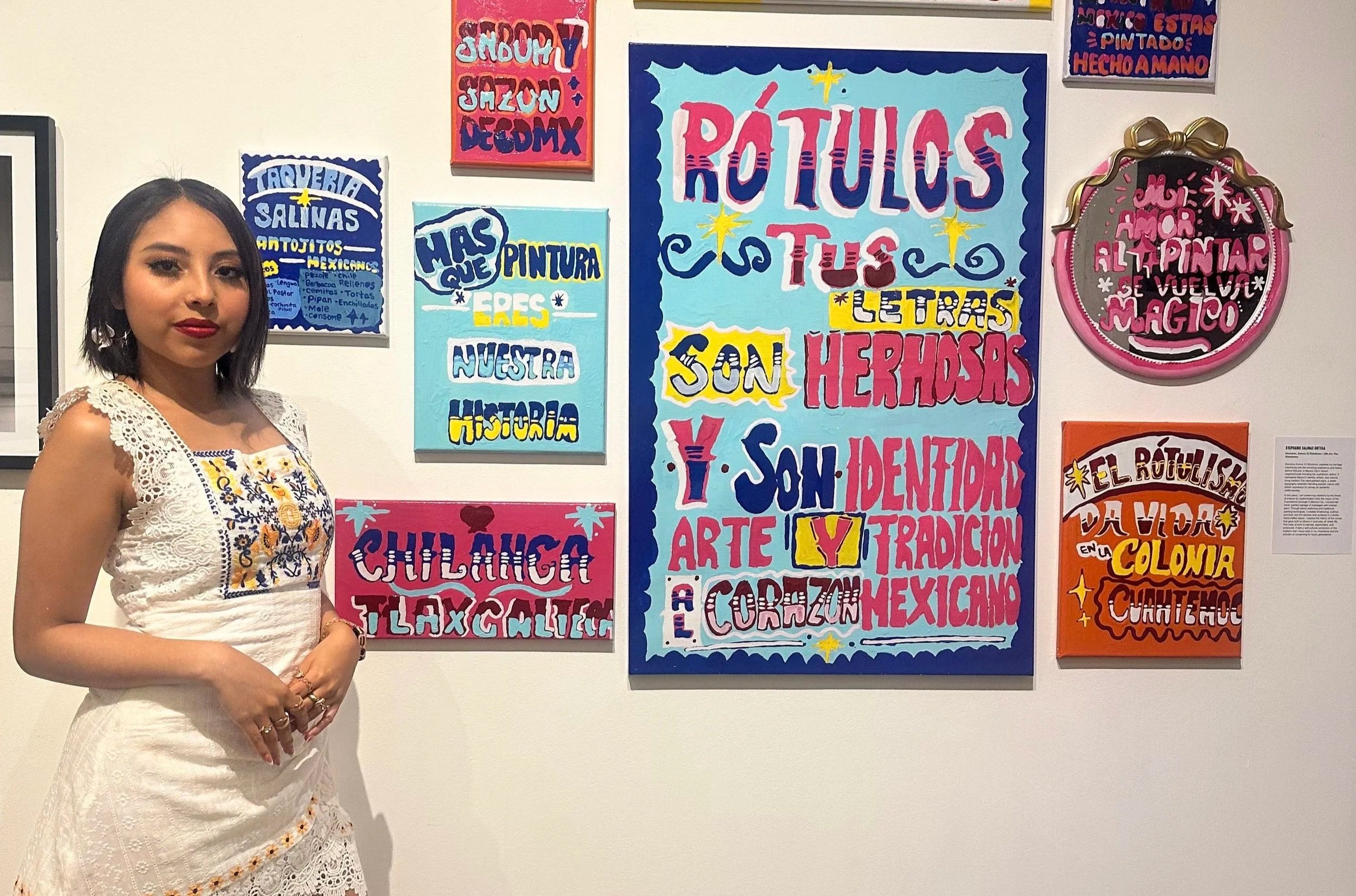 A young woman with short dark hair and red lipstick wearing a white embroidered dress, standing in front of colorful wall art in Spanish.