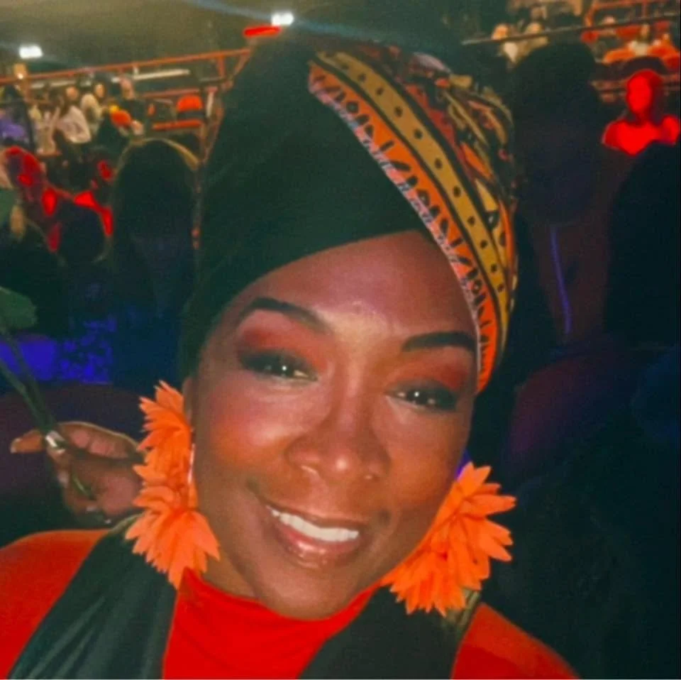A woman smiling, wearing orange flower earrings, red lipstick, and a colorful headwrap with yellow, black, and orange patterns. She is at a crowded outdoor event at night.