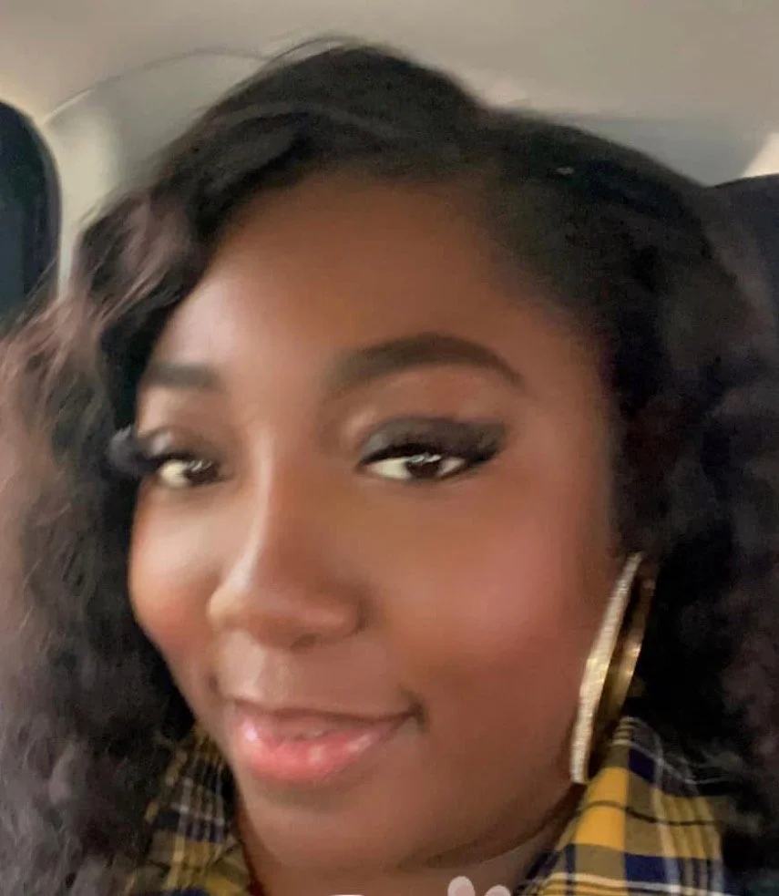 Close-up of a woman with curly black hair, wearing hoop earrings and a yellow plaid shirt, smiling softly.