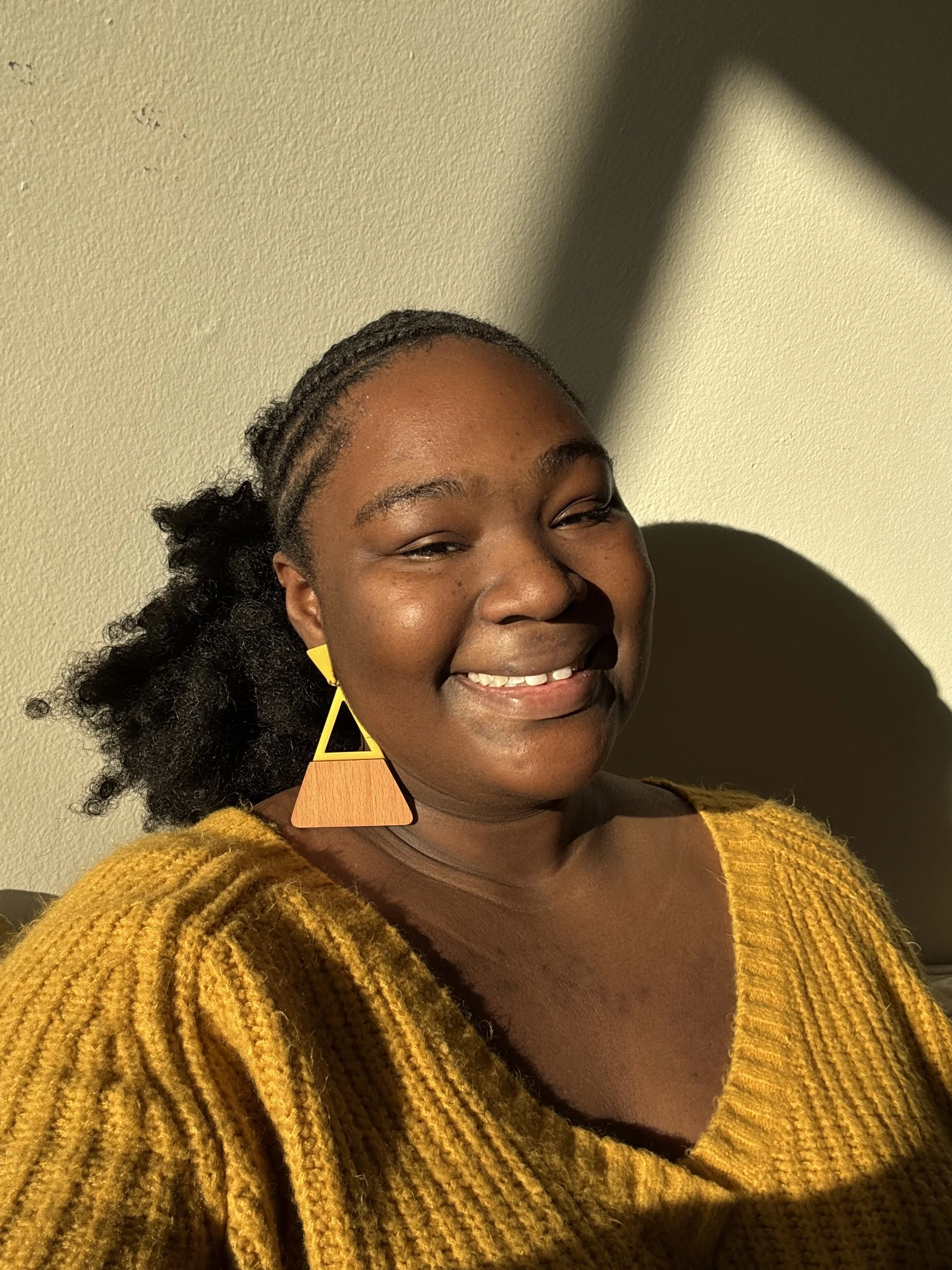 A smiling woman with dark skin and braided hair wearing large geometric earrings and a yellow knit sweater, standing against a plain wall.