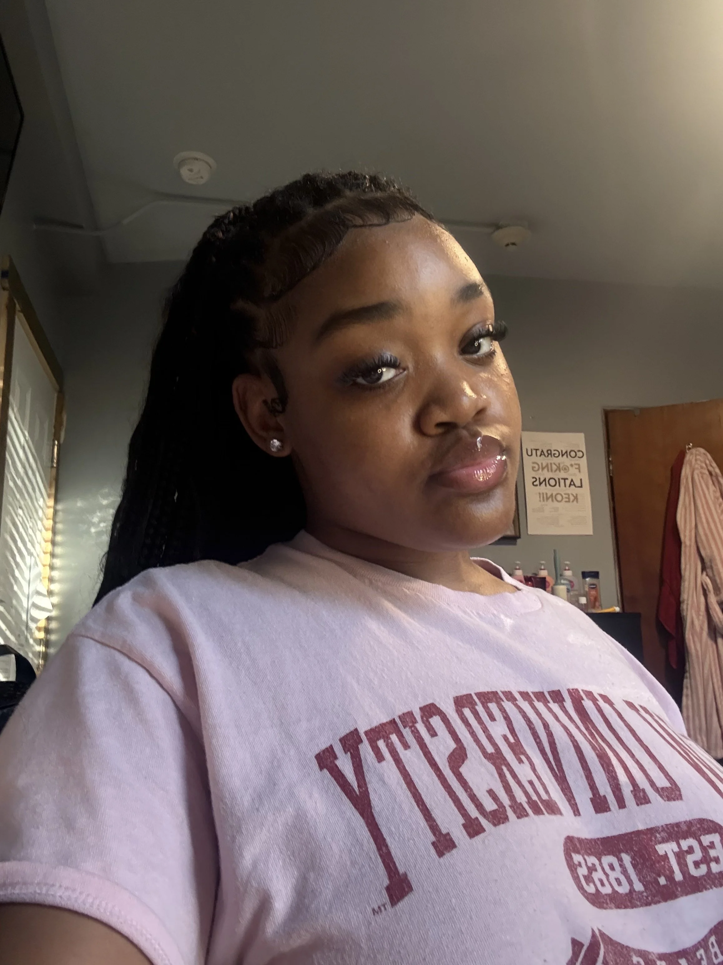 Close-up of a young woman's face, showing her eyes, lips, earrings, and curly braided hairstyle, with her wearing a pink T-shirt, in an indoor setting.