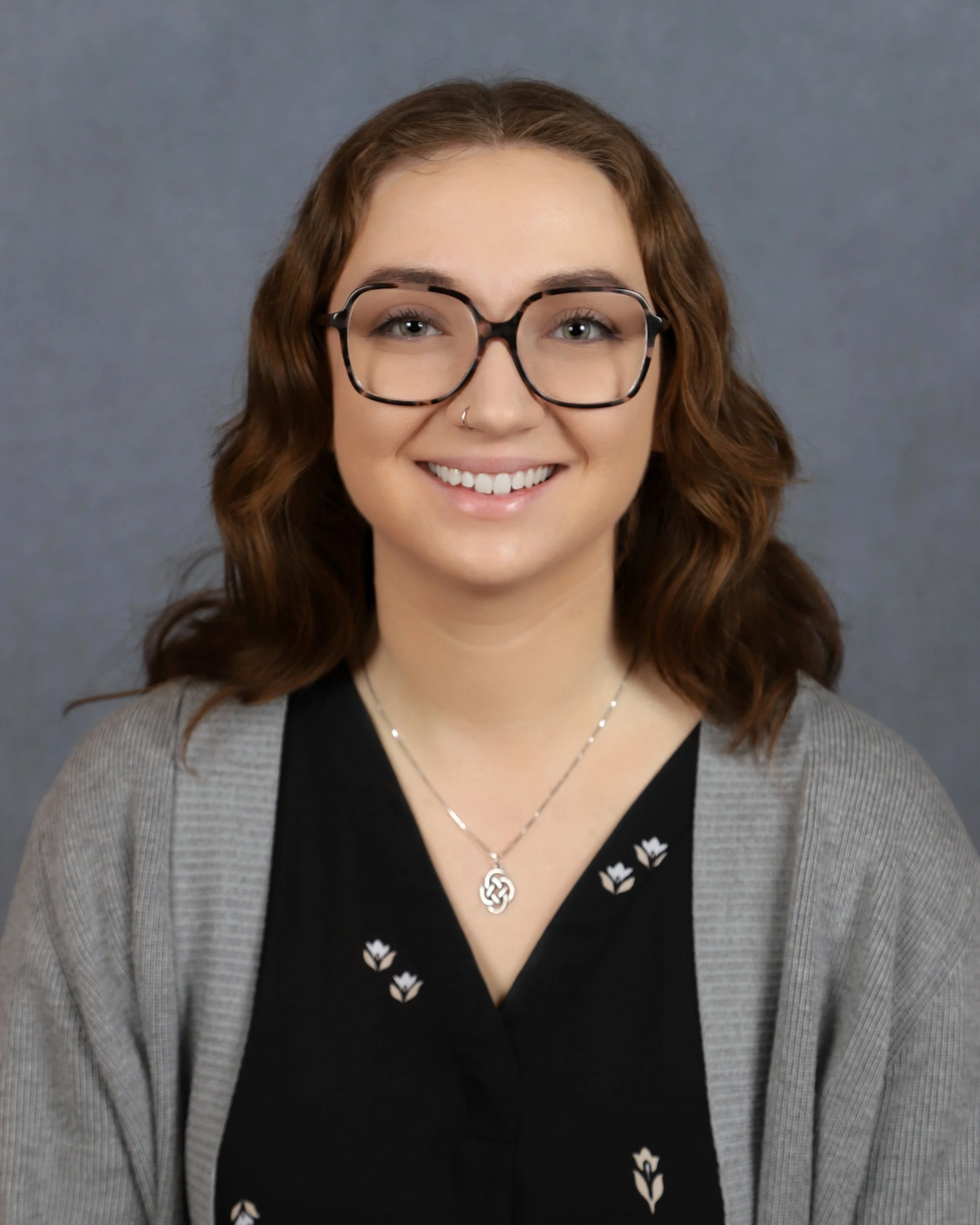 A woman with brown, wavy hair wearing glasses, a necklace, a nose ring, and a patterned black and gray top stands against a gray background, smiling.