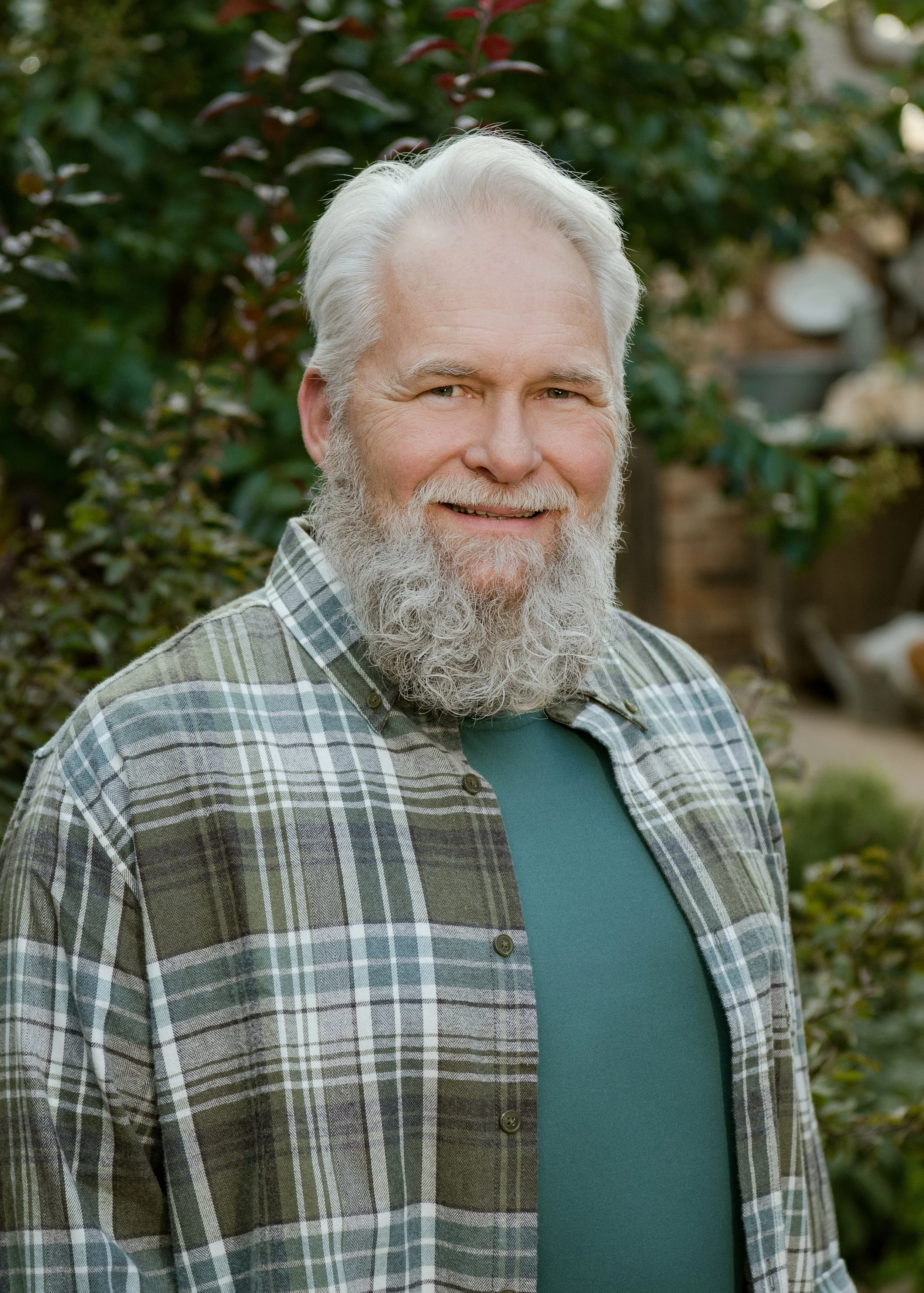 An elderly man with gray hair and a white beard smiling outdoors, wearing a green shirt and a plaid button-up shirt, with greenery in the background.