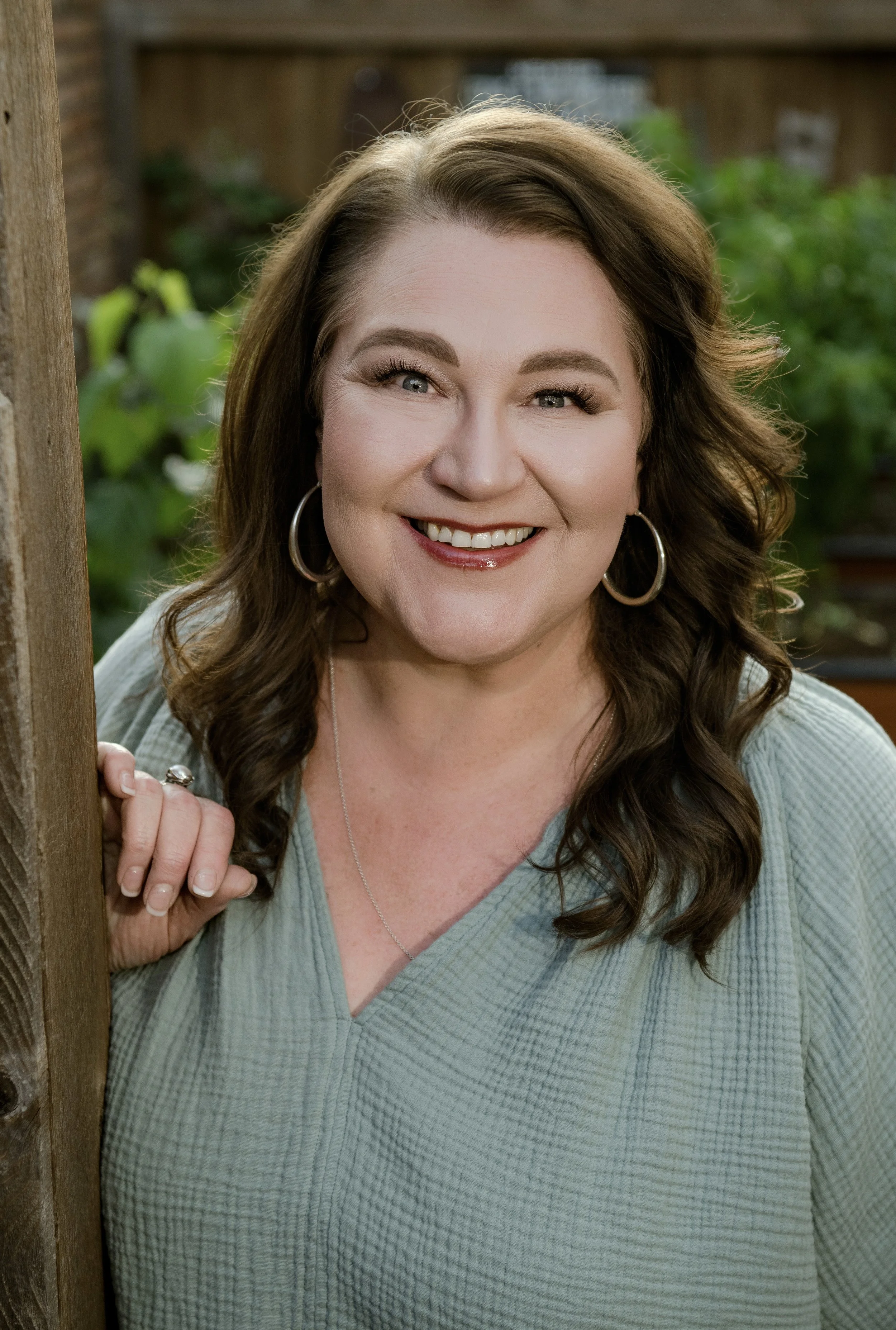 A woman with long wavy brown hair and hoop earrings smiling, peeking out from behind a wooden structure with a green background.