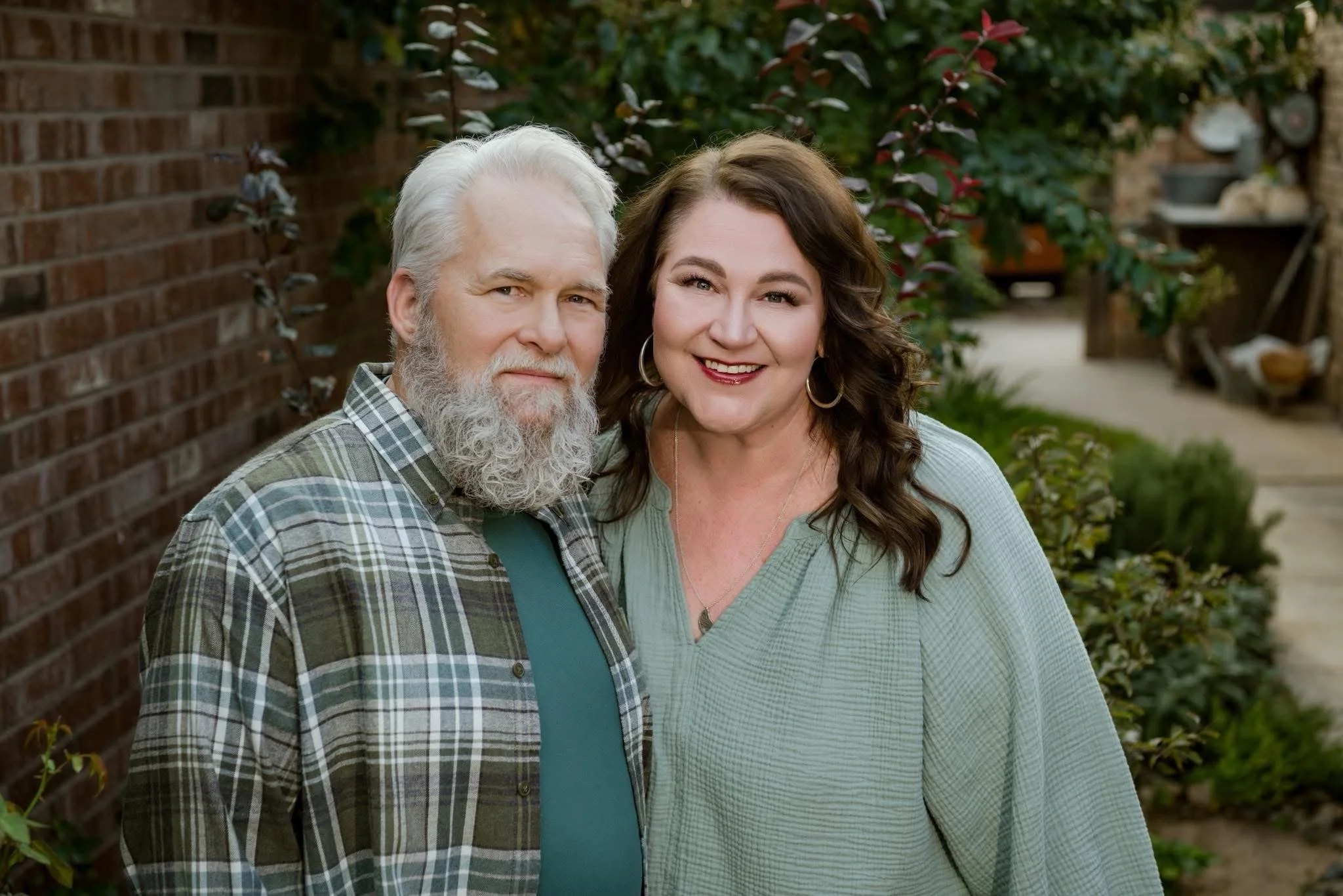 A middle-aged man with a white beard and gray hair and a middle-aged woman with brown curly hair, both smiling, standing outdoors near a brick wall and green shrubbery.