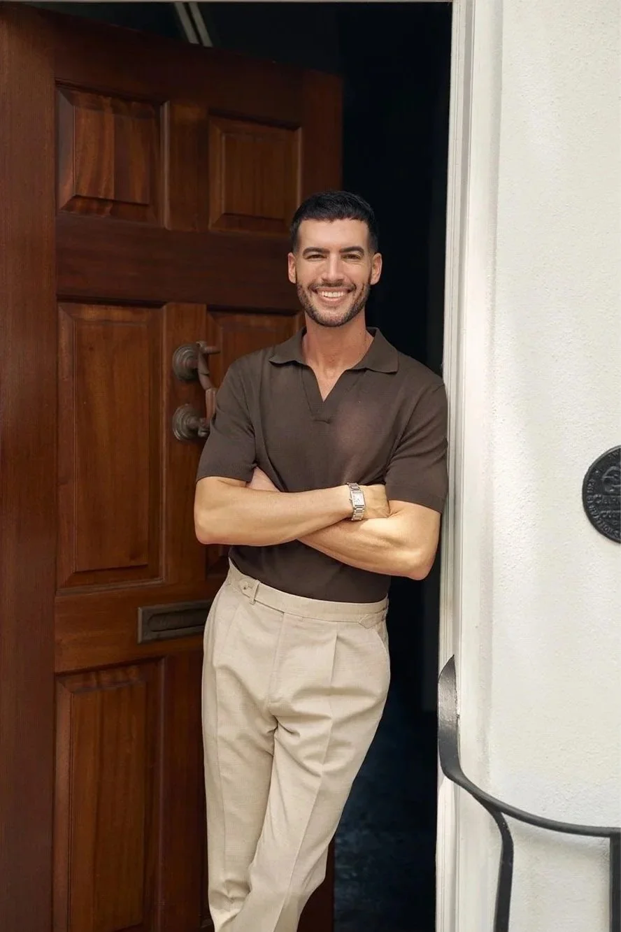 A man with dark hair and a beard standing in a doorway, smiling with arms crossed, wearing a brown polo shirt and beige trousers.