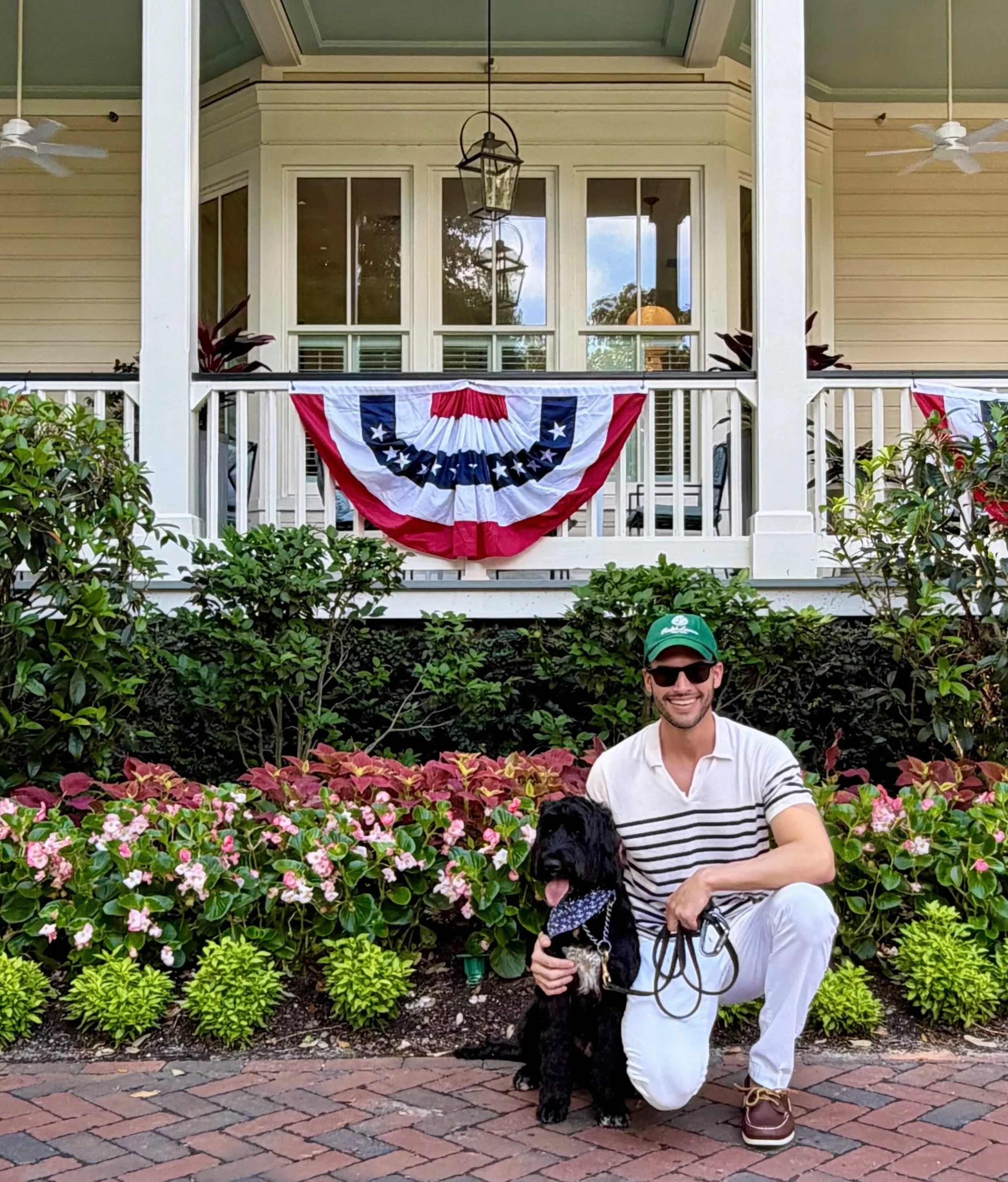 Ryan standing on a flower-filled porch with his black-and-white dog, who wears a bandana.