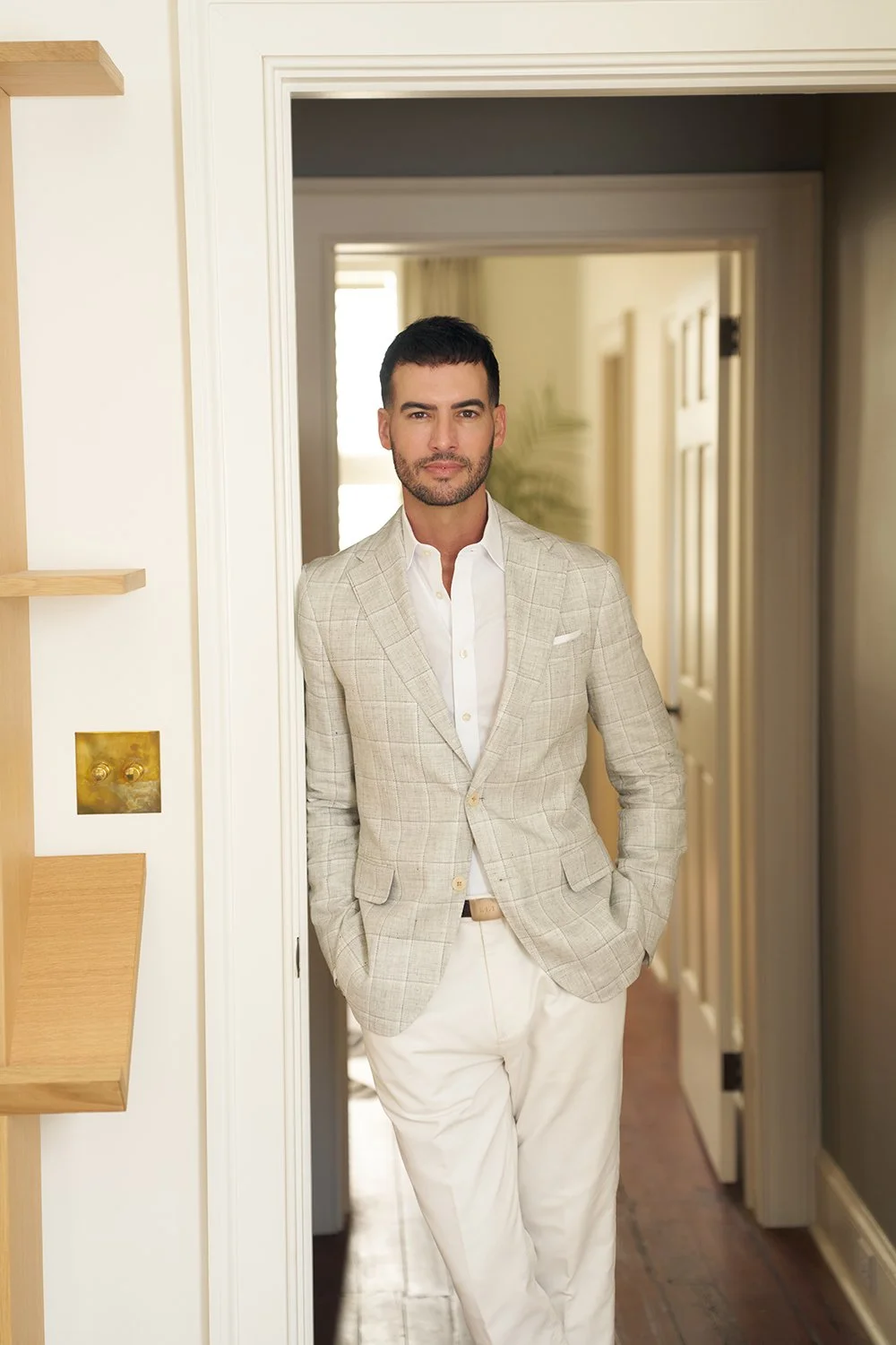 Ryan standing in a doorway in a beige blazer and white pants, one hand resting in his pocket.