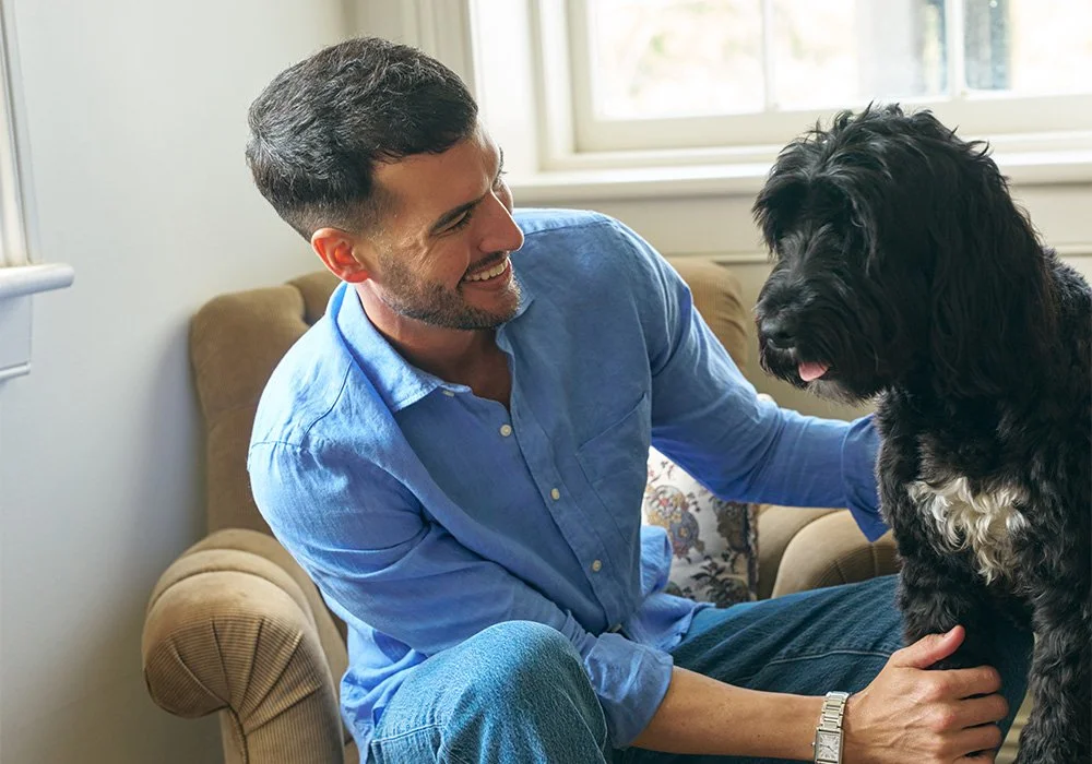 Ryan smiling at his black-and-white dog while sitting together, wearing a blue button-down shirt.