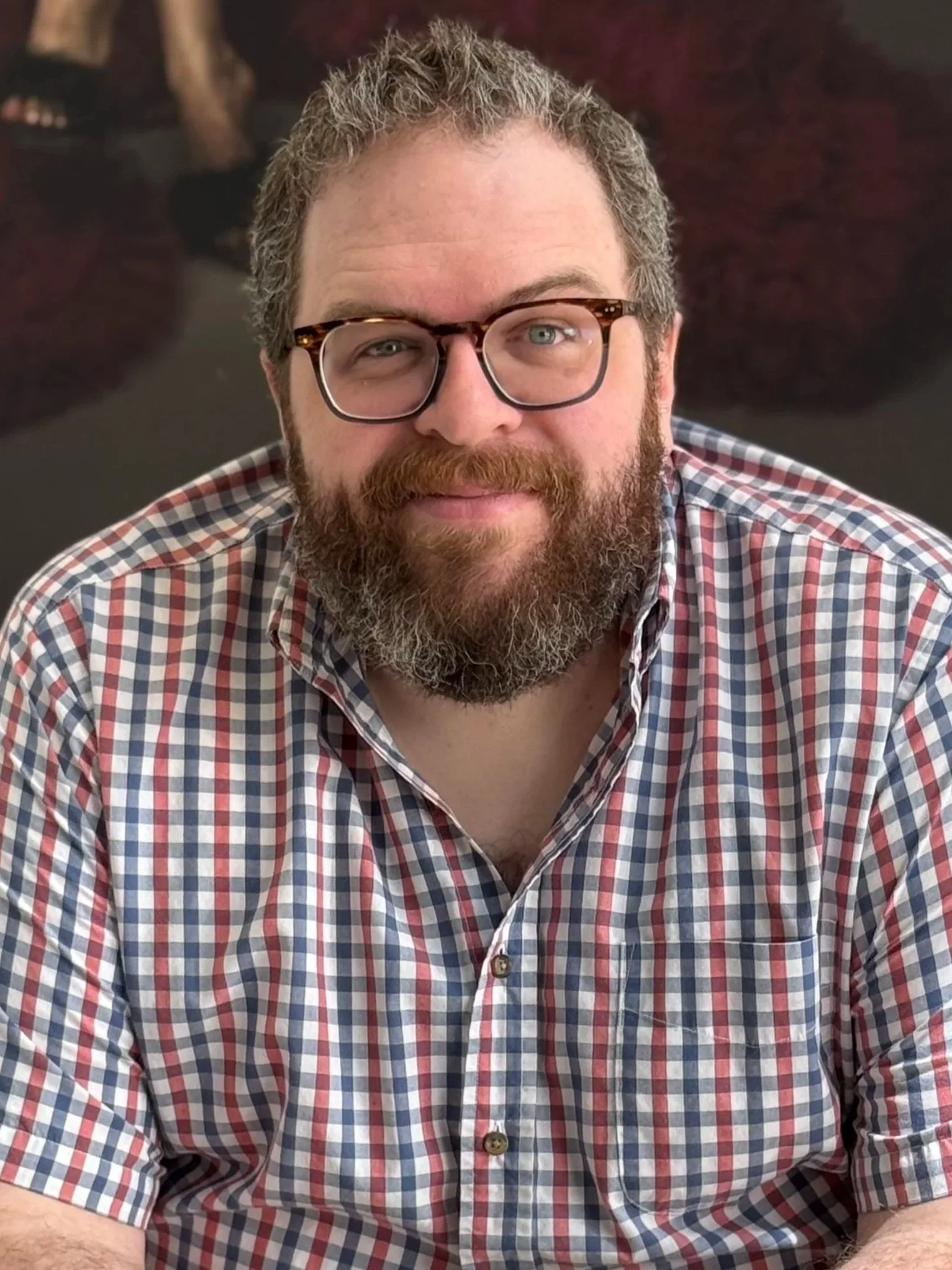 A bearded man with glasses and blue eyes wearing a red, white, and blue checkered shirt, smiling, with a dark background.