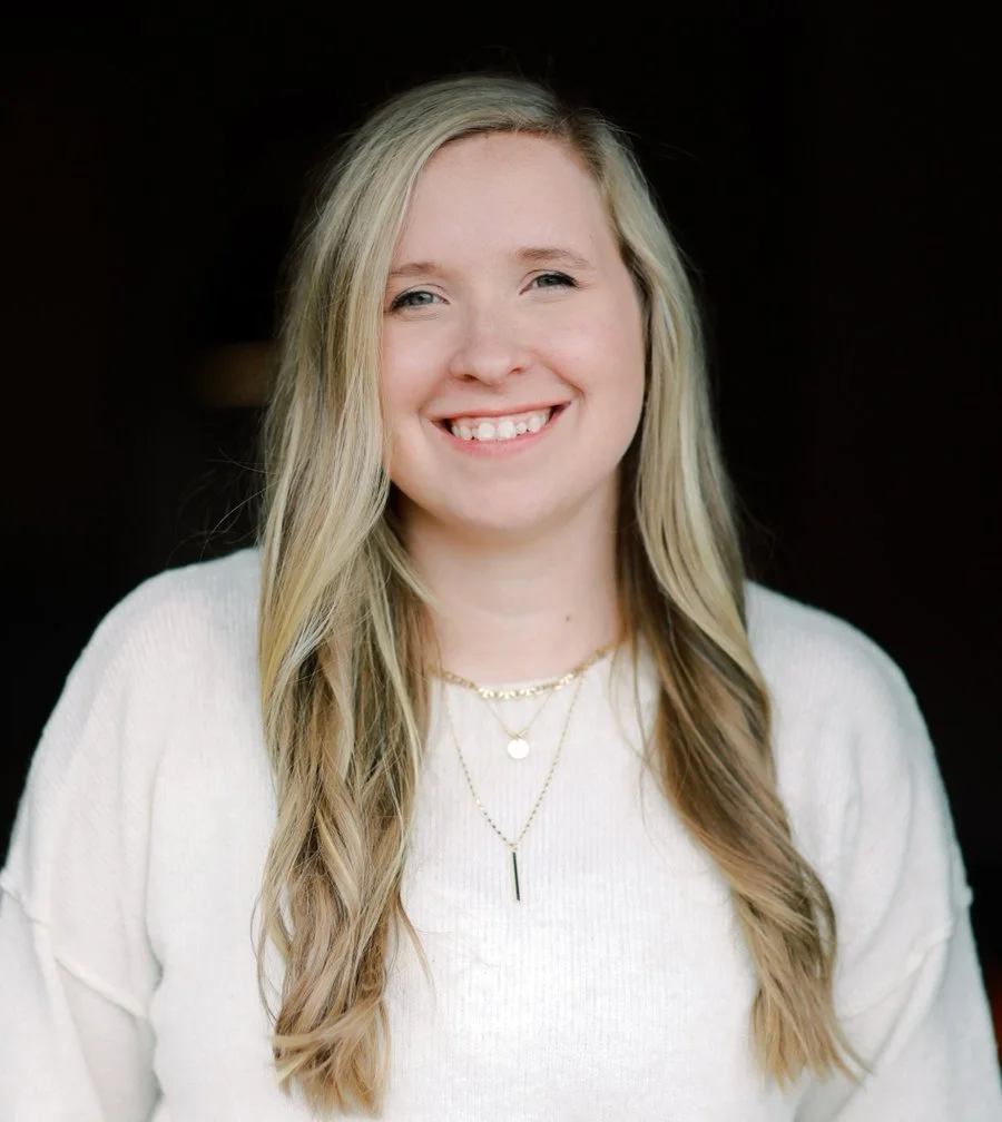 A young woman with long blonde hair smiling against a dark background, wearing a white sweater and layered gold necklaces.