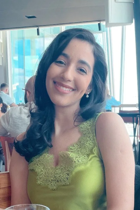 A young woman with dark, wavy hair and pearl earrings, smiling, wearing a lime green dress with lace detailing, sitting in a restaurant with large windows and other patrons in the background.