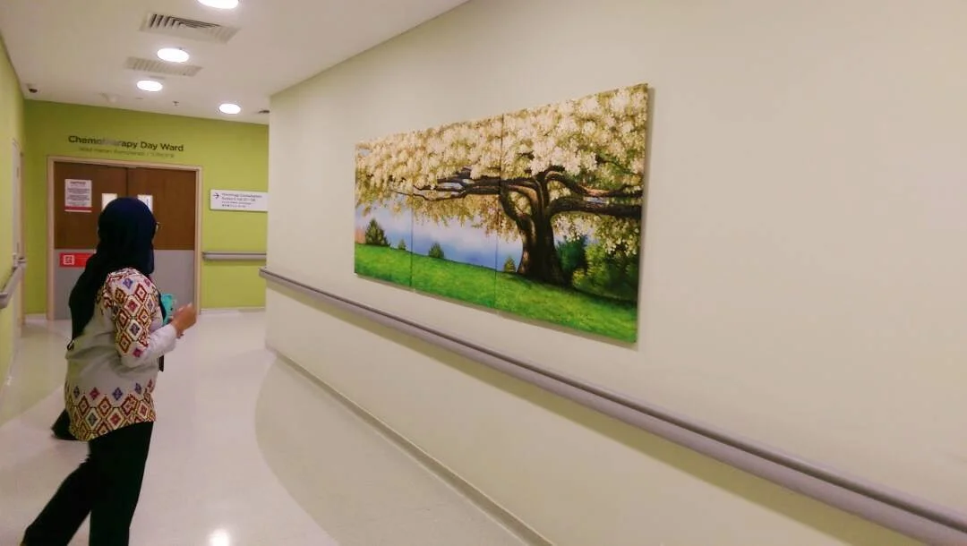 A woman in a patterned jacket and headscarf looking at a painting of a blossoming tree on a hospital corridor wall.