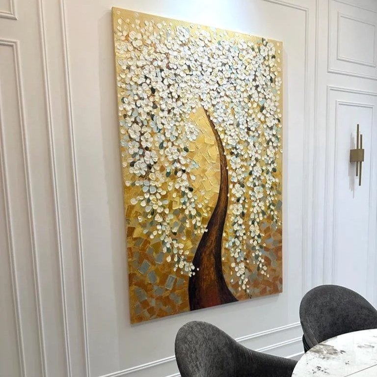 A large, textured painting of a tree with white blossoms on a gold background, displayed on a white wall in a modern interior space.
