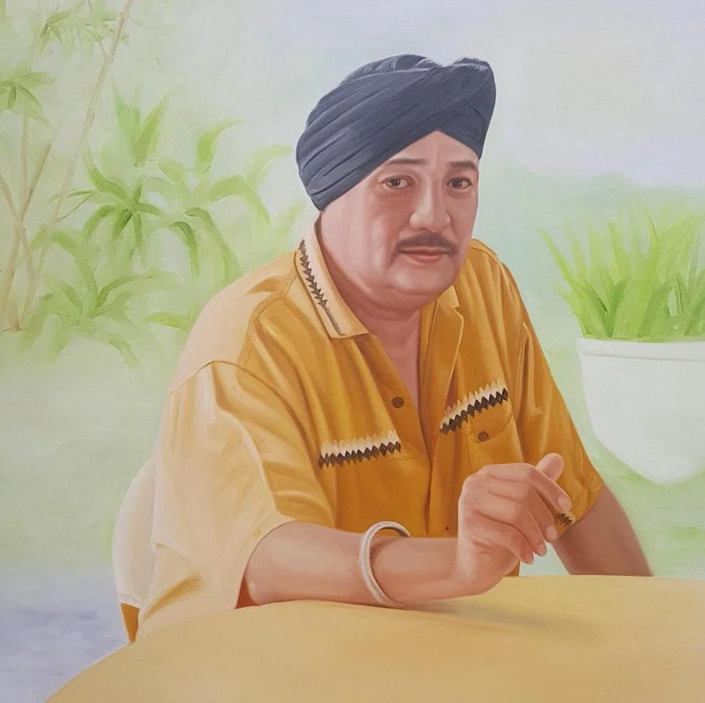 A man wearing a black turban and a mustard yellow shirt with black and white zigzag patterns sitting at a yellow table with a light green background, which includes green plants in pots.