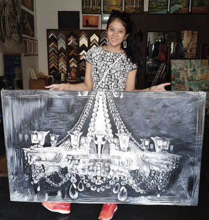 Young woman holding a large black and white painting of a chandelier with candles and jewelry, standing in an art gallery.