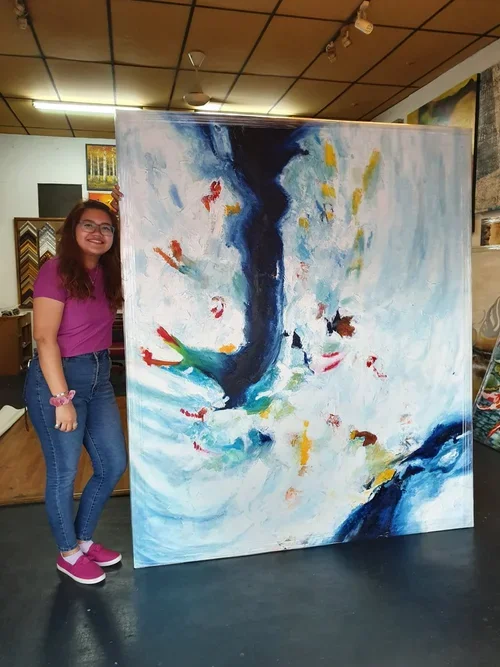 A young woman in a pink shirt and jeans stands next to a large abstract painting in an art studio. The painting features bold strokes of blue, white, and spots of red, green, and yellow, with a central dark blue shape.