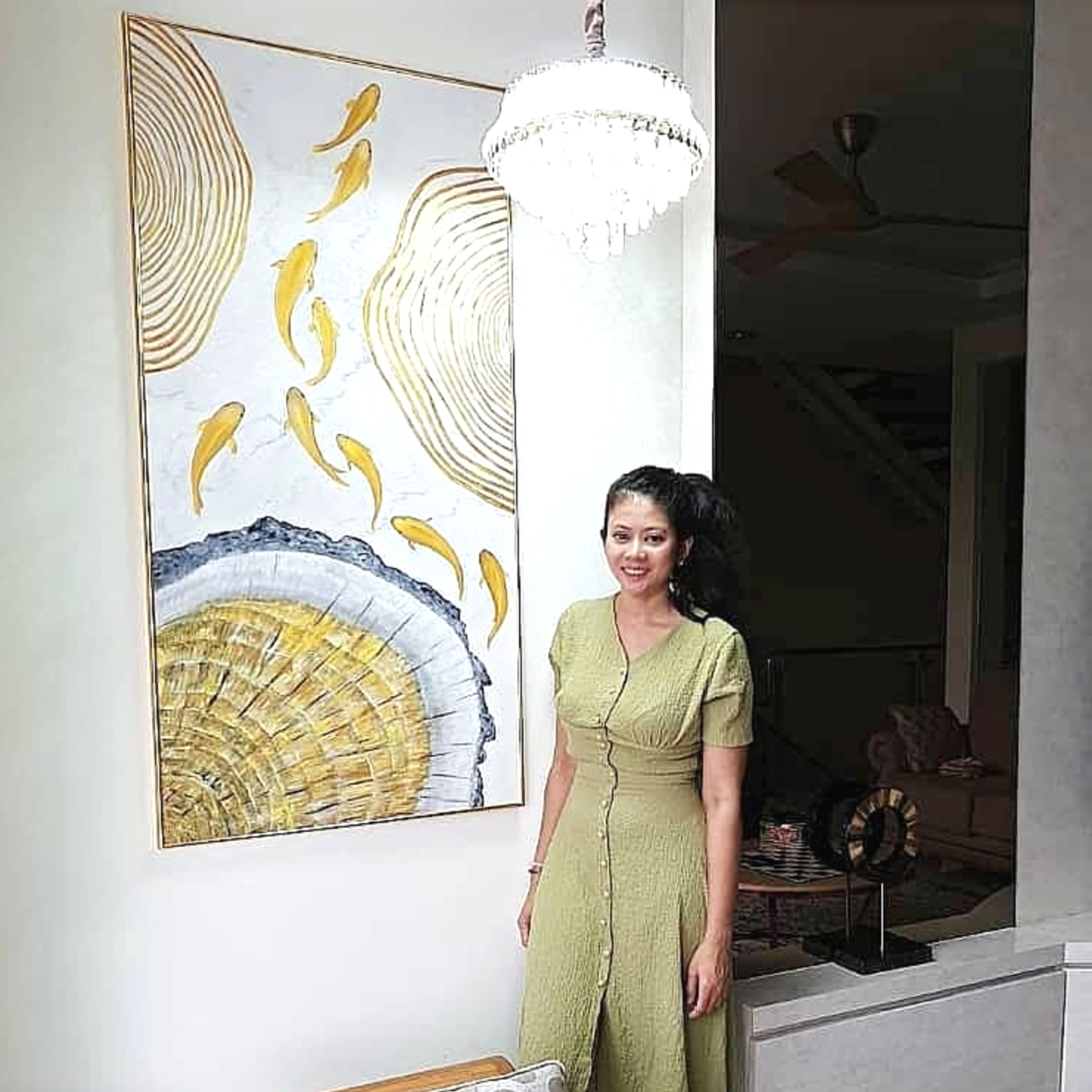 A woman standing next to a large abstract painting of a mountain and yellow fish, with a chandelier hanging from the ceiling and a ceiling fan in the background.
