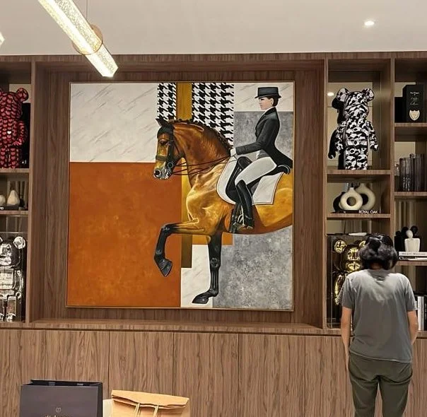 A person observing a large painting of a rider on a horse, with the rider dressed in equestrian attire and the horse in mid-gallop, mounted on a wooden wall with shelves displaying decorative items.