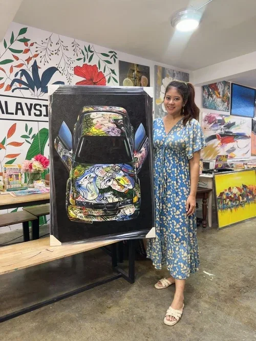 A woman is standing next to a large painting of a car with abstract colorful designs inside it. The woman is wearing a blue and yellow floral dress and smiling. The background shows a decorated wall with various artworks and a table with flowers and 