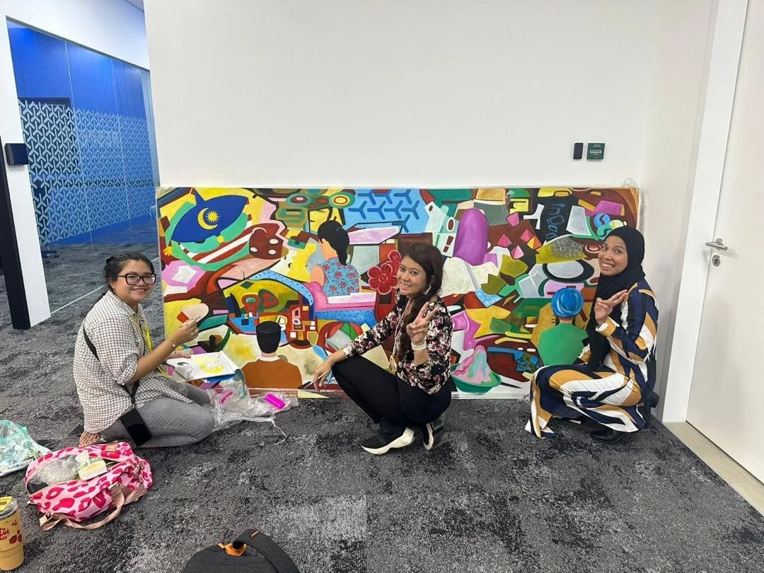 Three women sitting on the floor in front of a colorful abstract mural, smiling and posing for the photo. One woman is wearing glasses and checkered clothing, another is in a floral top, and the third is wearing a patterned hijab and striped outfit.