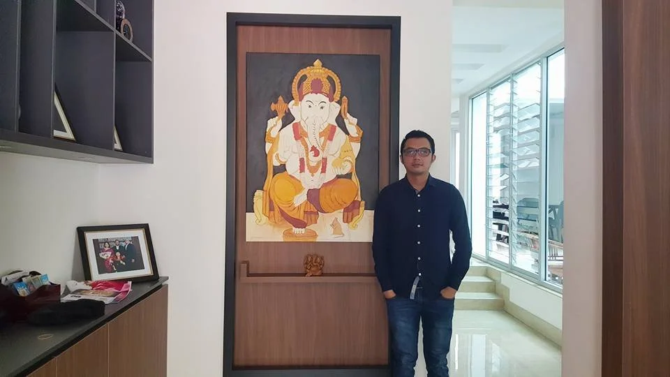 A man standing next to a framed painting of the Hindu deity Ganesha in an indoor space with a modern design. The background features a large window with blinds and an outside view.