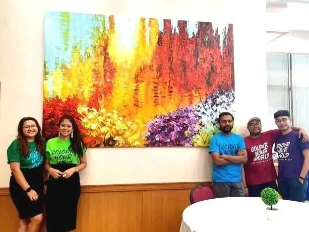 Group of five people standing indoors in front of a large, colorful abstract painting, with a round table and small green plant in the foreground.