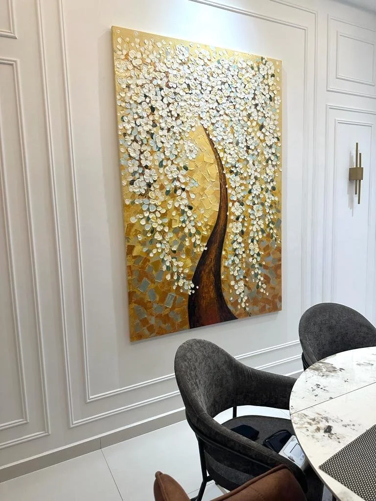 A painting of a stylized tree with a tall, slender trunk and white blossoms set against a gold background, hanging on a white paneled wall in a modern interior space.