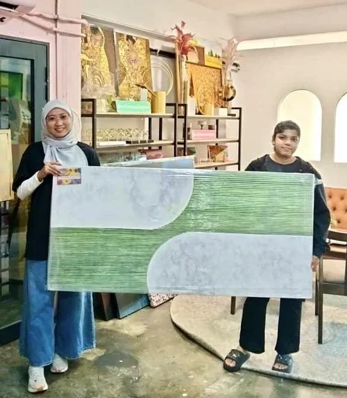 Two women holding a large rectangular artwork with green and white patterns in an art studio with shelves of art supplies and decorations.