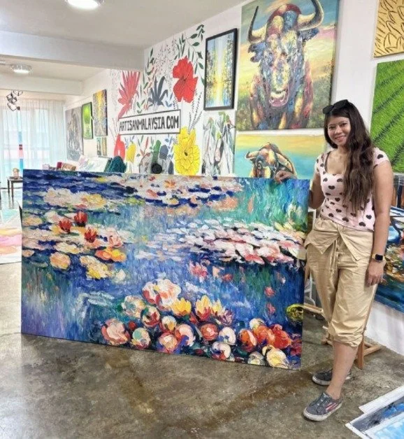 A woman standing indoors next to a large colorful painting of lotus flowers on water, with various artworks hanging on the wall behind her, including a vibrant cow portrait and floral designs.