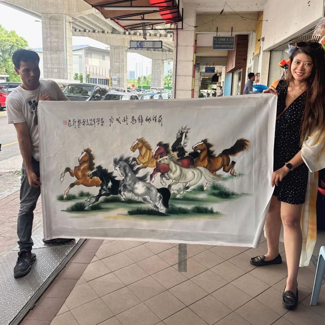 Two people holding a large fabric scroll with a painting of ten galloping horses in traditional Asian style, under a covered outdoor area near parked cars and a highway overpass.