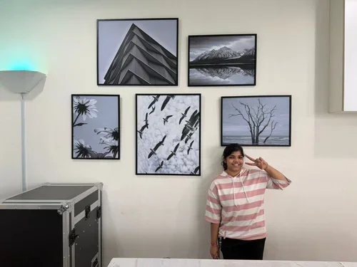 A woman standing in front of a gallery wall with five framed landscape photos, making a peace sign. The photos include architecture, nature, and trees.