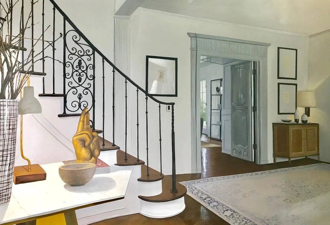 Interior of a house with a staircase, decorative iron balusters, framed artwork on the walls, a doorway leading to another room, a side table with vases and a lamp, and a large area rug on hardwood floor.