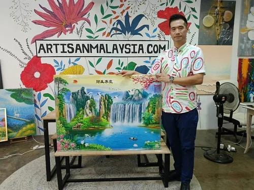 A man wearing a colorful shirt stands next to a landscape painting of waterfalls and lush greenery, displayed on an easel inside an art studio. There are other paintings, a fan, and a colorful floral mural with the website ARTISANMALAYSIA.COM on the 