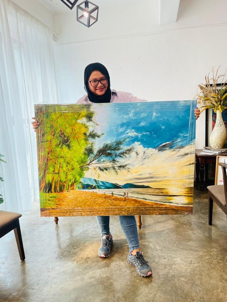 A woman wearing glasses and a black hijab holding a large landscape painting of a beach with trees, mountains, and a cloudy sky, inside a room with white walls, sheer curtains, and a ceiling light fixture.