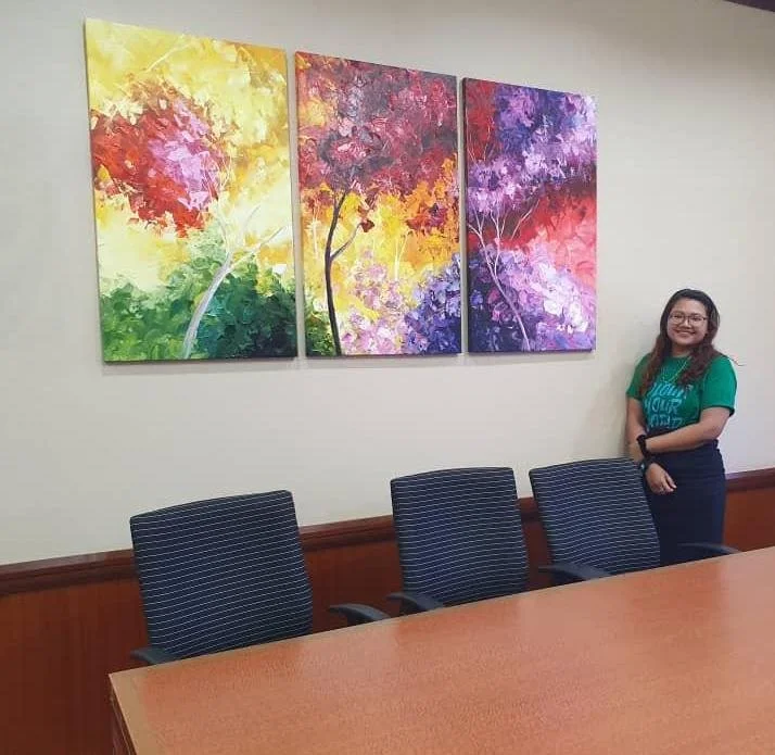 A woman standing in a conference room next to three colorful abstract paintings of trees on a white wall.