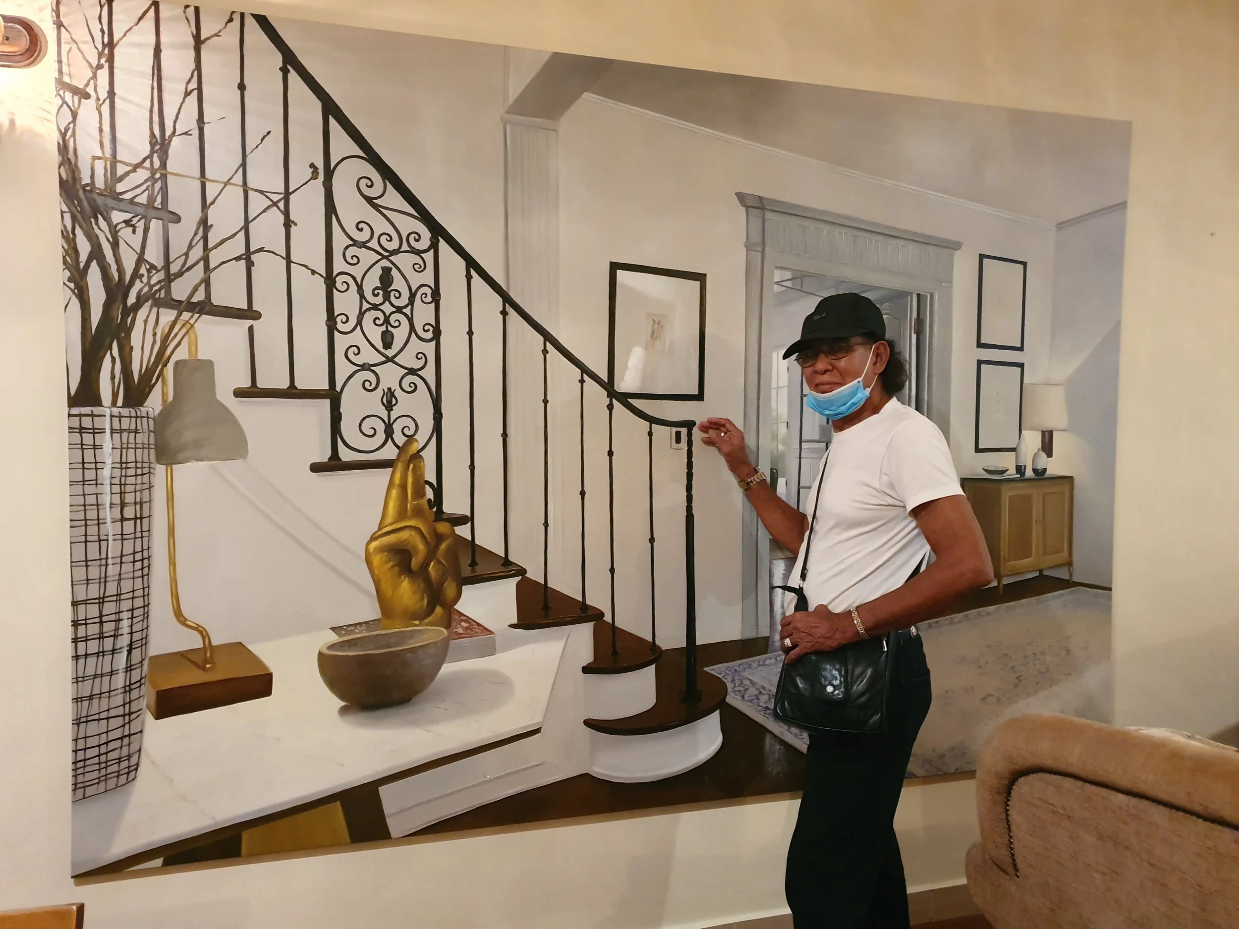 An older woman with glasses and a face mask, wearing a white shirt and black hat, stands in a home decor store next to a table with decorative objects, including a golden hand sculpture, vases, and a spiral staircase with ornate black iron railing.