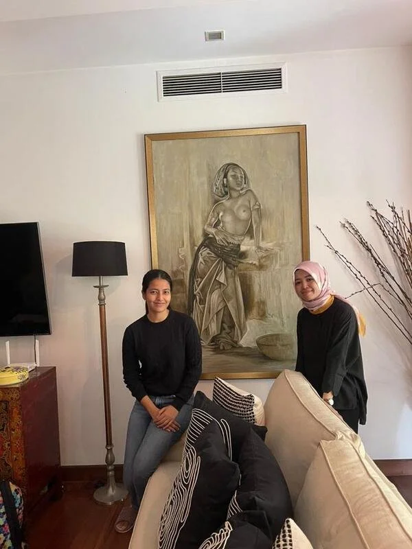 Two women standing in a living room, one with dark hair in a black sweater and jeans, the other wearing a pink headscarf and black top. Behind them, a large artwork of a seated woman with a headscarf, and a standing lamp. There are cushions on a beig